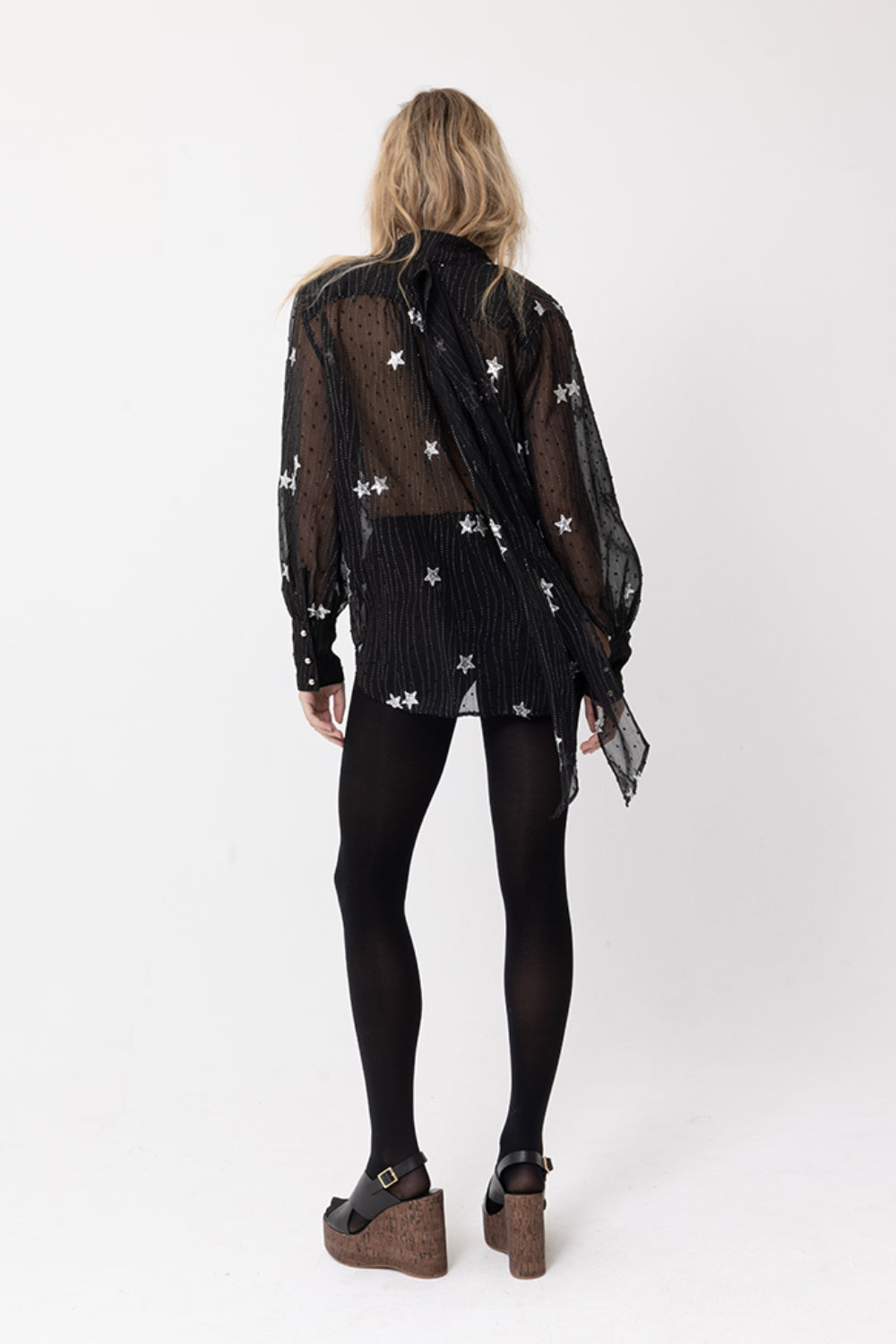 Back view of model wearing JOEY the Label sheer black long-sleeve blouse with silver sequin star embellishment, ruffle v-neck and neck-tie. Styled with black stockings.