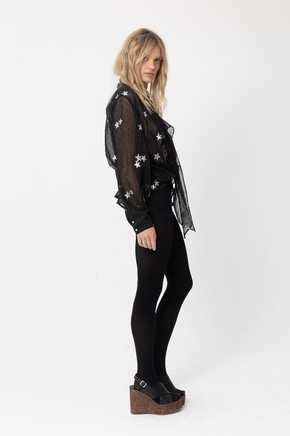 Side view of model wearing JOEY the Label sheer black long-sleeve blouse with silver sequin star embellishment, ruffle v-neck and neck-tie. Styled with black stockings.