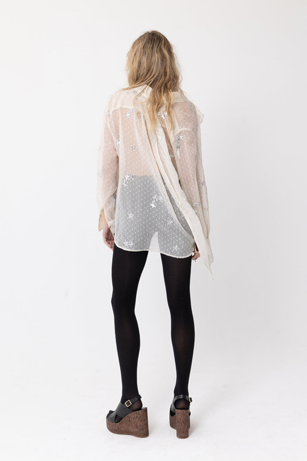 Back view of model wearing JOEY the Label sheer beige long-sleeve blouse with silver sequin star embellishments, ruffle v-neck and neck-tie. Styled with black stockings.