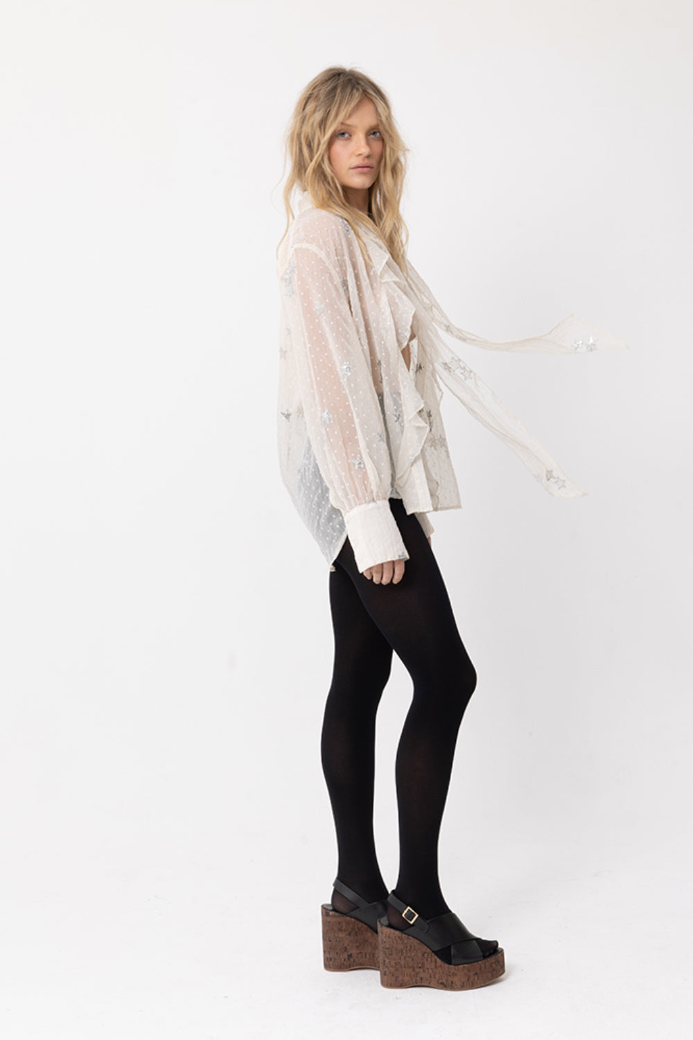 Side view of model wearing JOEY the Label sheer beige long-sleeve blouse with silver sequin star embellishments, ruffle v-neck and neck-tie. Styled with black stockings.